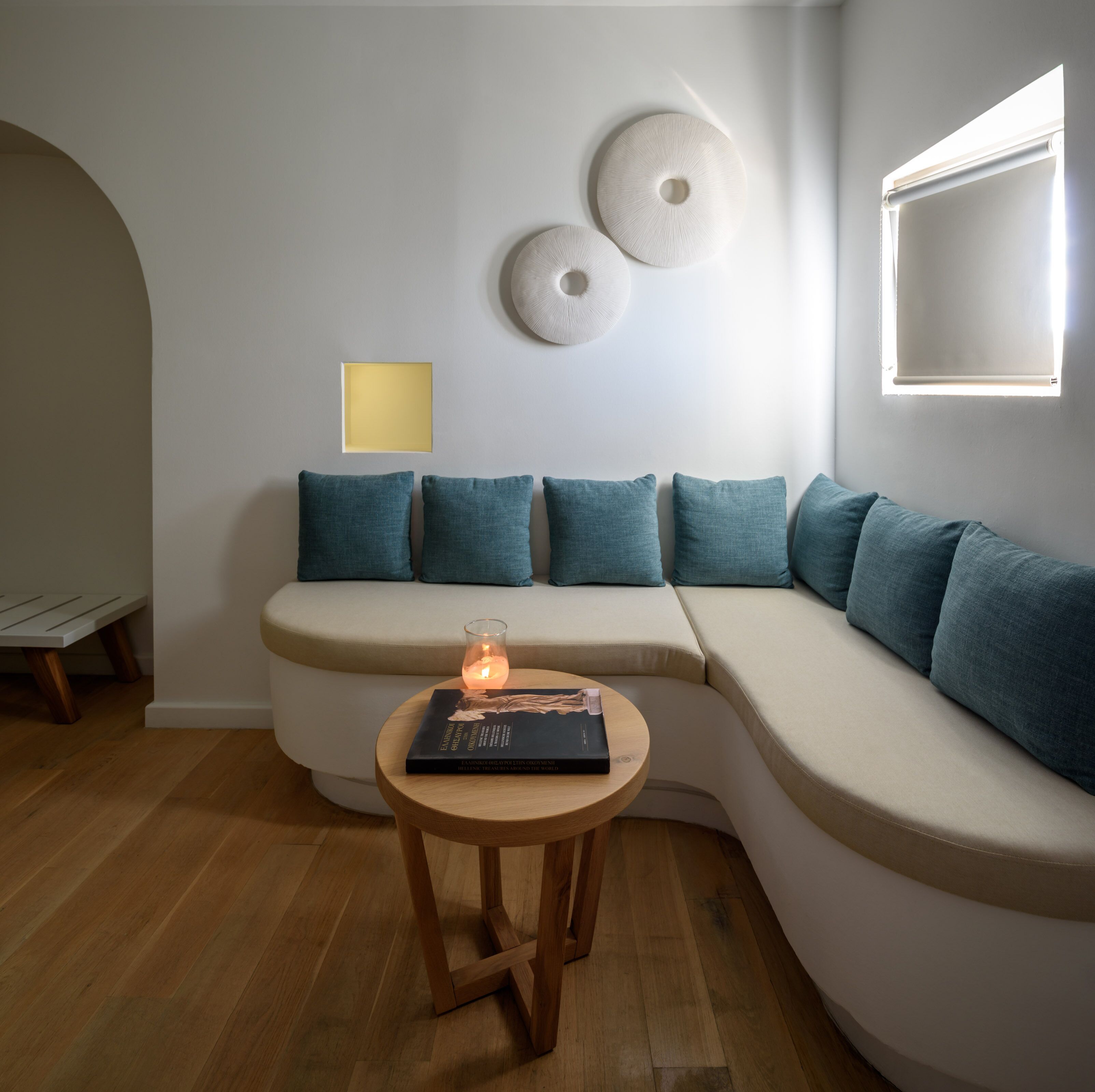 Ein modernes Wohnzimmer mit einer gebogenen Couch, Kissen in Türkis, einem kleinen Holztisch mit einem Buch und einer Kerze sowie einer Wand mit dekorativen Tellern.