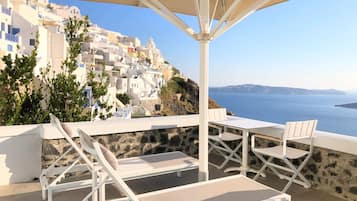 Une terrasse dotée de chaises longues blanches et d’une table, offrant une vue sur une ville côtière aux bâtiments blancs et sur la mer.
