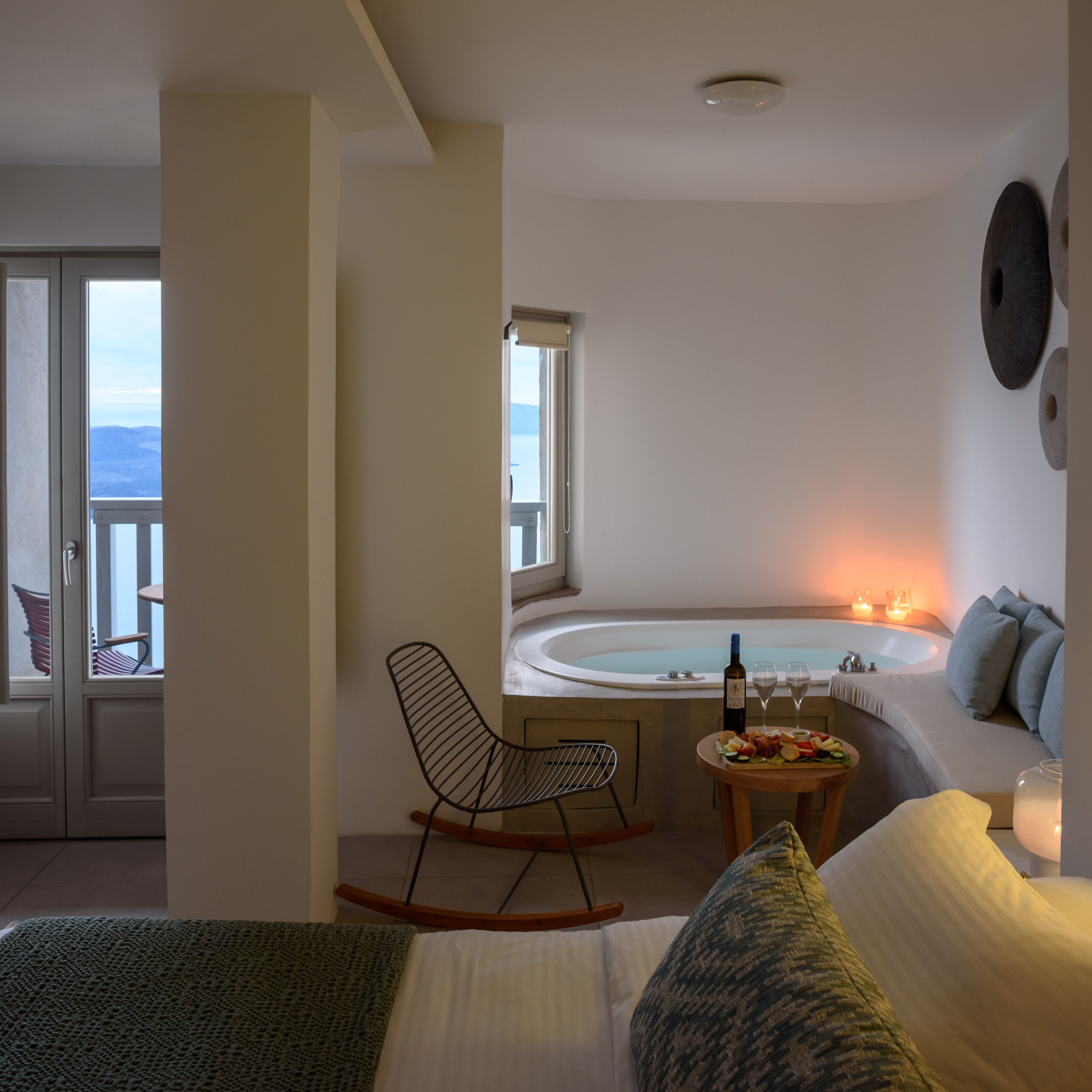 Un dormitorio moderno con una cama grande, una mecedora, una mesita con una botella y vasos, y una bañera con velas.