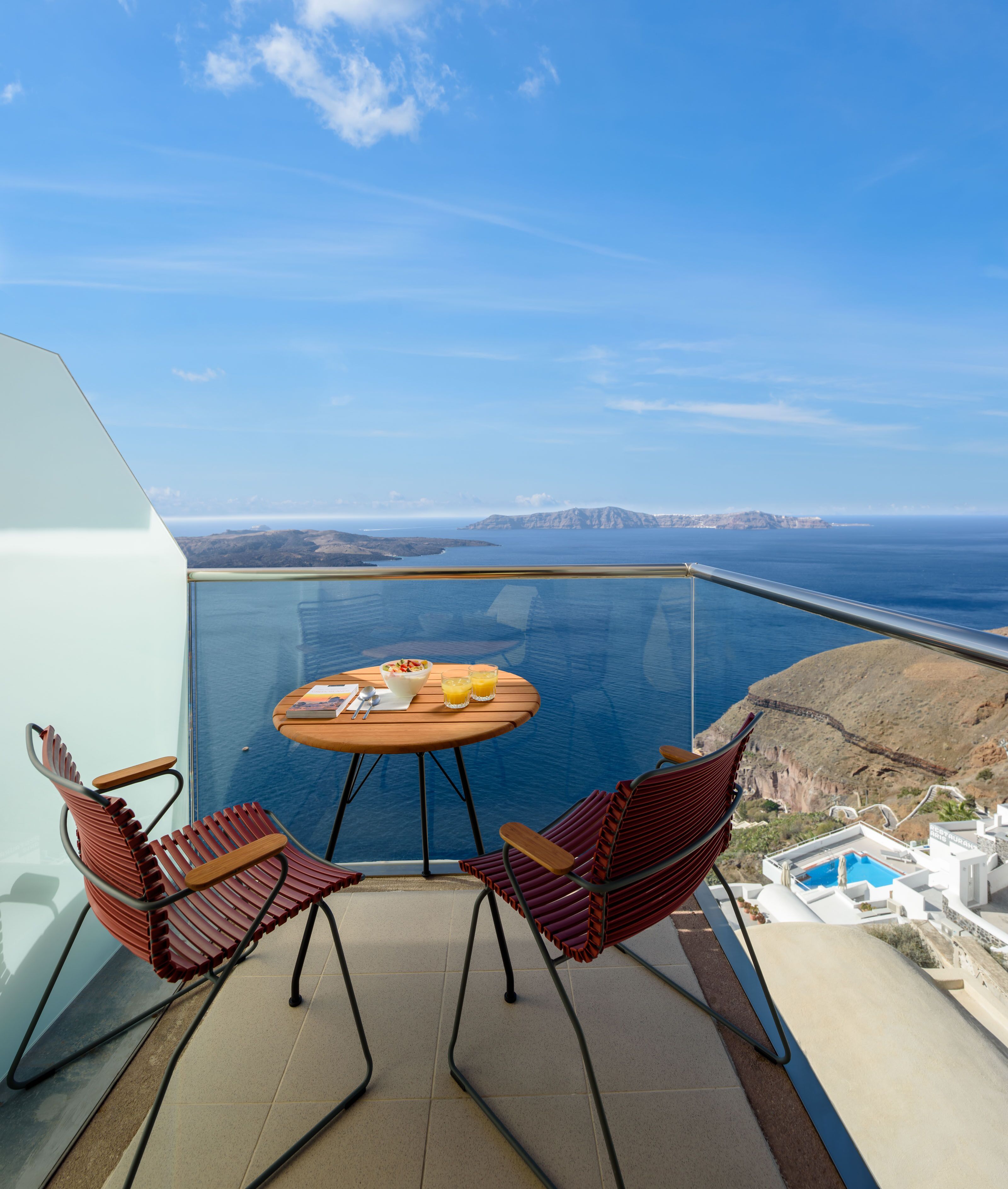 Een balkon met glazen rand en uitzicht op de kust, met een tafel, stoelen en een ontbijtje.