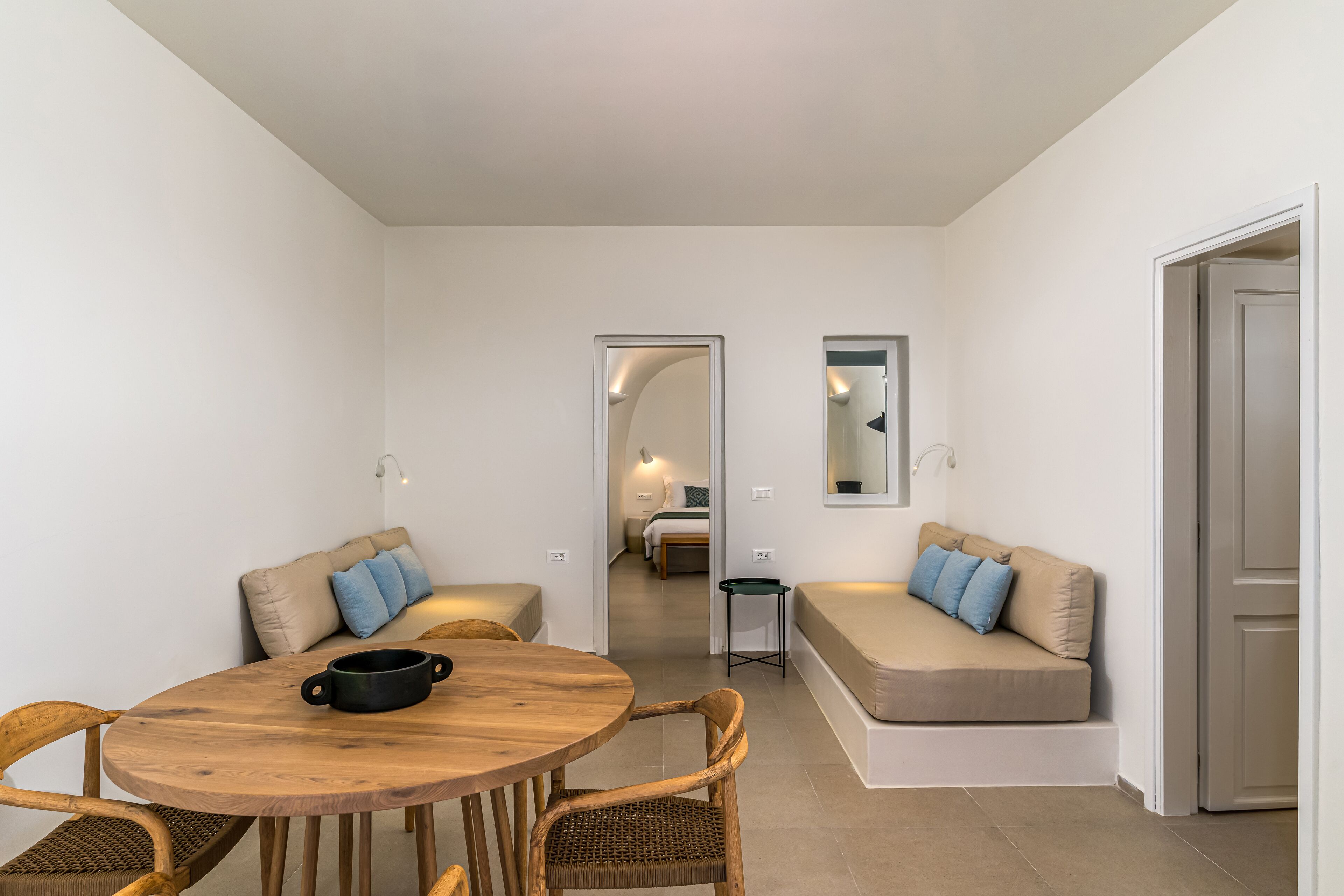 A compact living space with a round wooden table, two cushioned seating areas, a small side table, and a doorway leading to a bedroom.