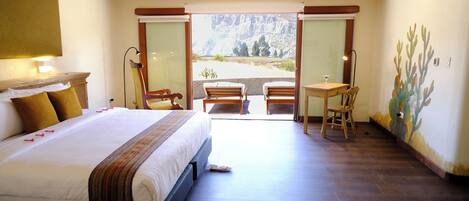 Una habitación de hotel con una cama grande, un balcón con vistas a las montañas, una mesita con una silla y un cuadro en la pared.