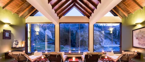 A spa room with wooden ceiling beams, large windows offering a view of a mountainous landscape, and multiple sun loungers with candles.