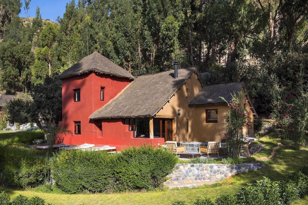 A red and beige house with a thatched roof, surrounded by a garden and forest.