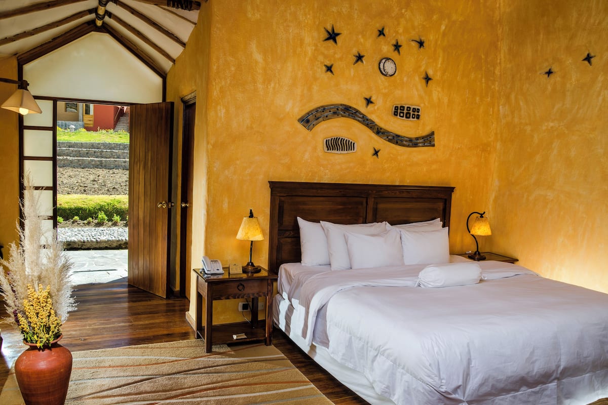 A hotel room with a bed, bedside lamps, a wooden nightstand, a vase with pampas grass, and a wall mural.