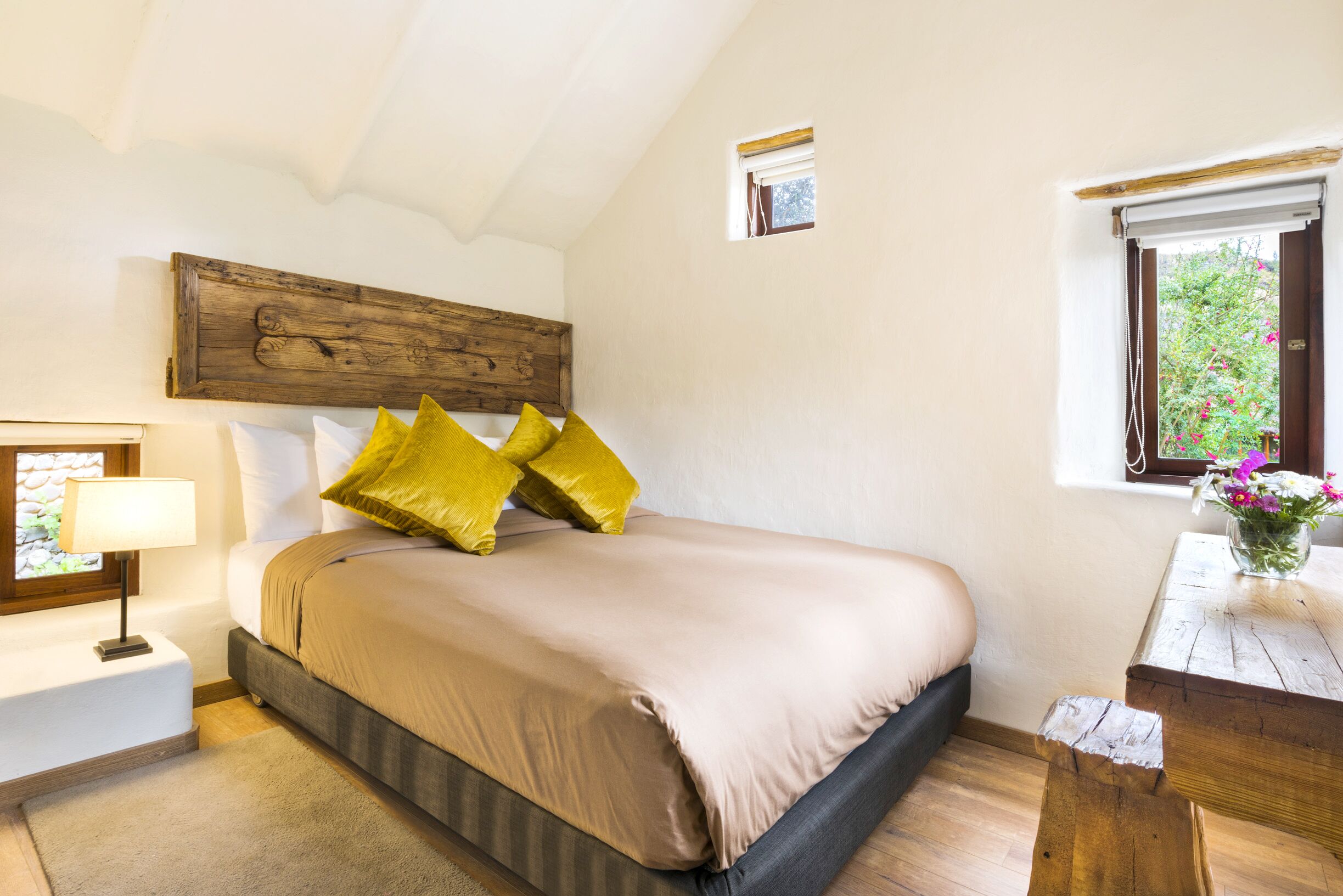 Un dormitorio con cama, mesita de noche, cabecera de madera, ventana con vistas a la vegetación y una pequeña mesa con un jarrón de flores.