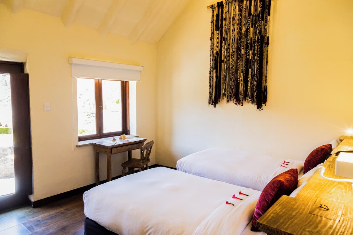 A bedroom with two beds, a wooden desk, and a window with blinds.