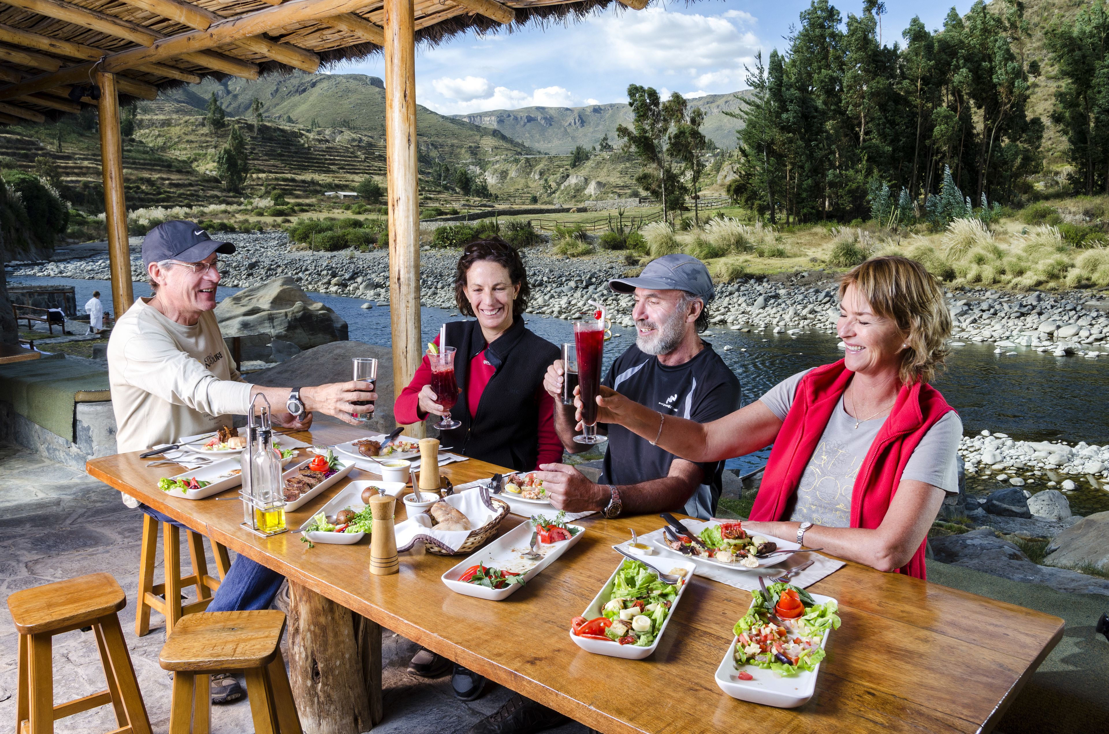 Una mesa de madera dispuesta con platos de comida, vasos y una botella, rodeada de gente en un entorno natural pintoresco.