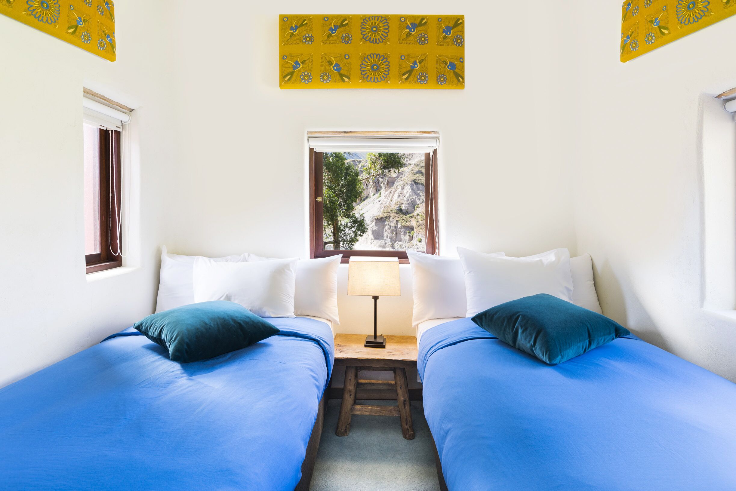 Una habitación con dos camas, una ventana que da a árboles y montañas, y una mesita con una lámpara.