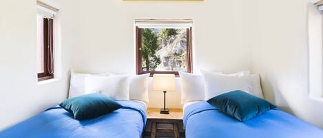 Una habitación con dos camas, una ventana que da a árboles y montañas, y una mesita con una lámpara.