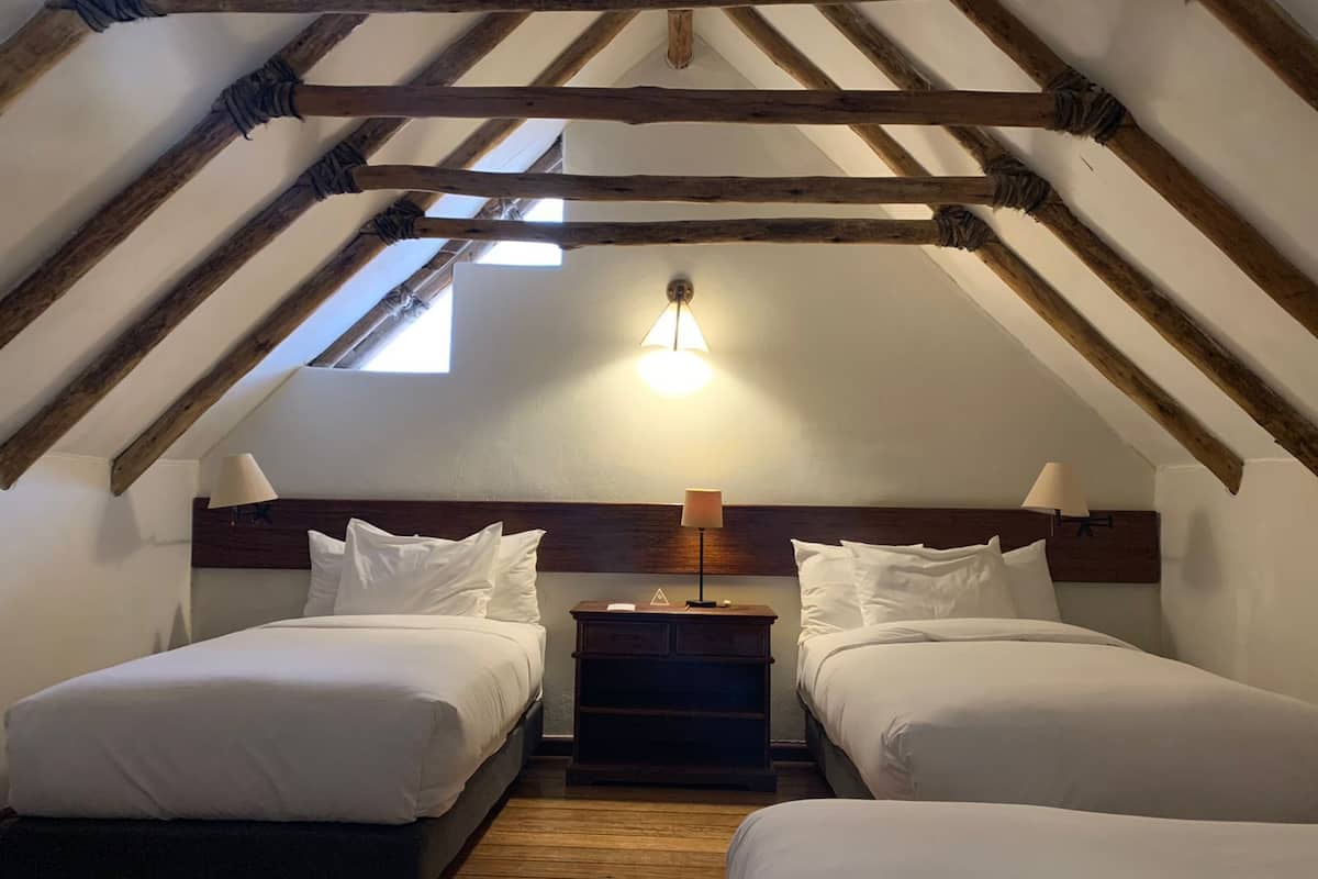 A room with two beds, wooden beams, a sloped ceiling, and a single light fixture.