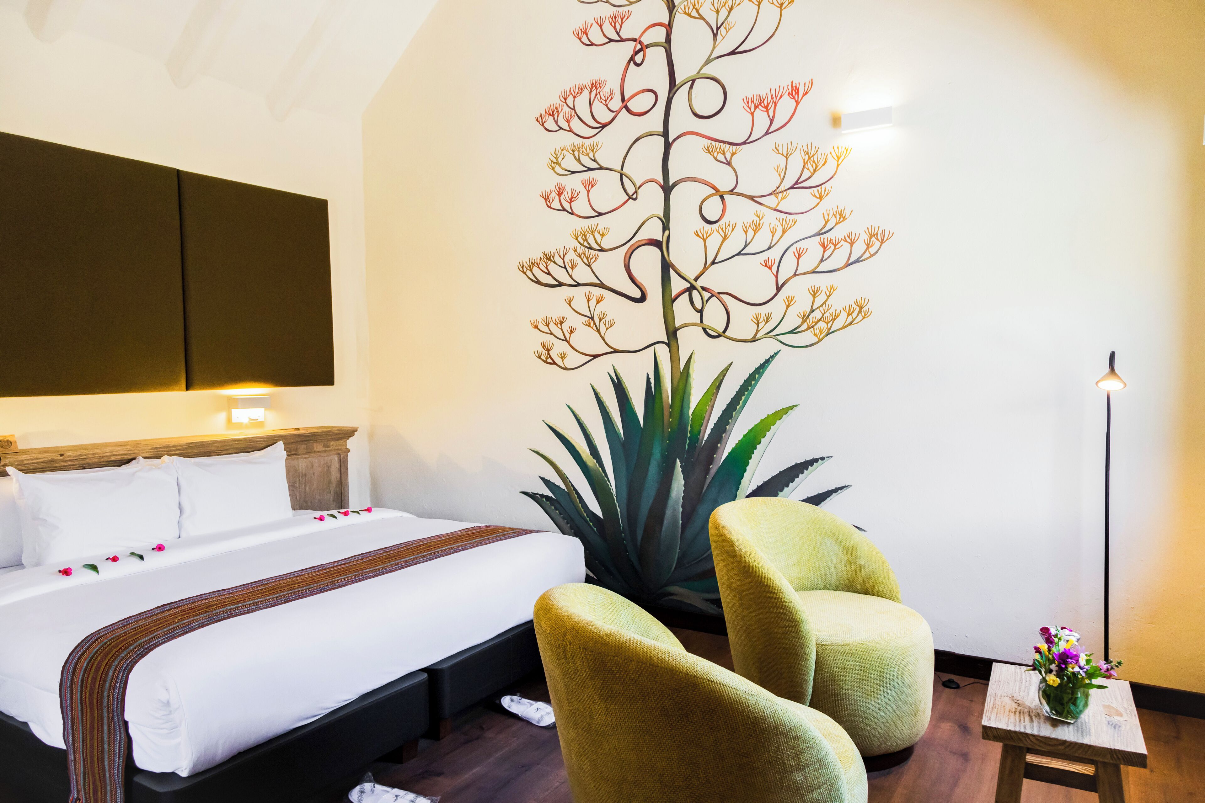 A hotel room with a large bed, two green armchairs, a small table with a vase of flowers, and a wall mural.
