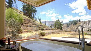 Un baño con una bañera grande, un ventanal con vistas a un paisaje montañoso y una mesita con una vela y un florero.