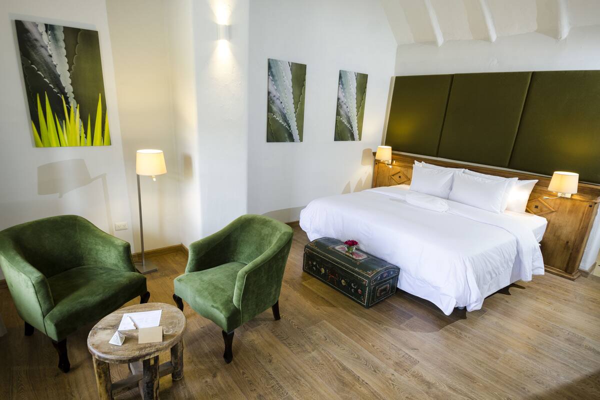 A hotel room with a large bed, two green armchairs, a wooden coffee table, and a vintage trunk.