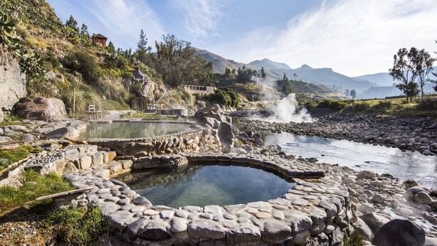 Un área de aguas termales naturales con una piscina de piedra, rodeada de rocas y vegetación, y montañas al fondo.