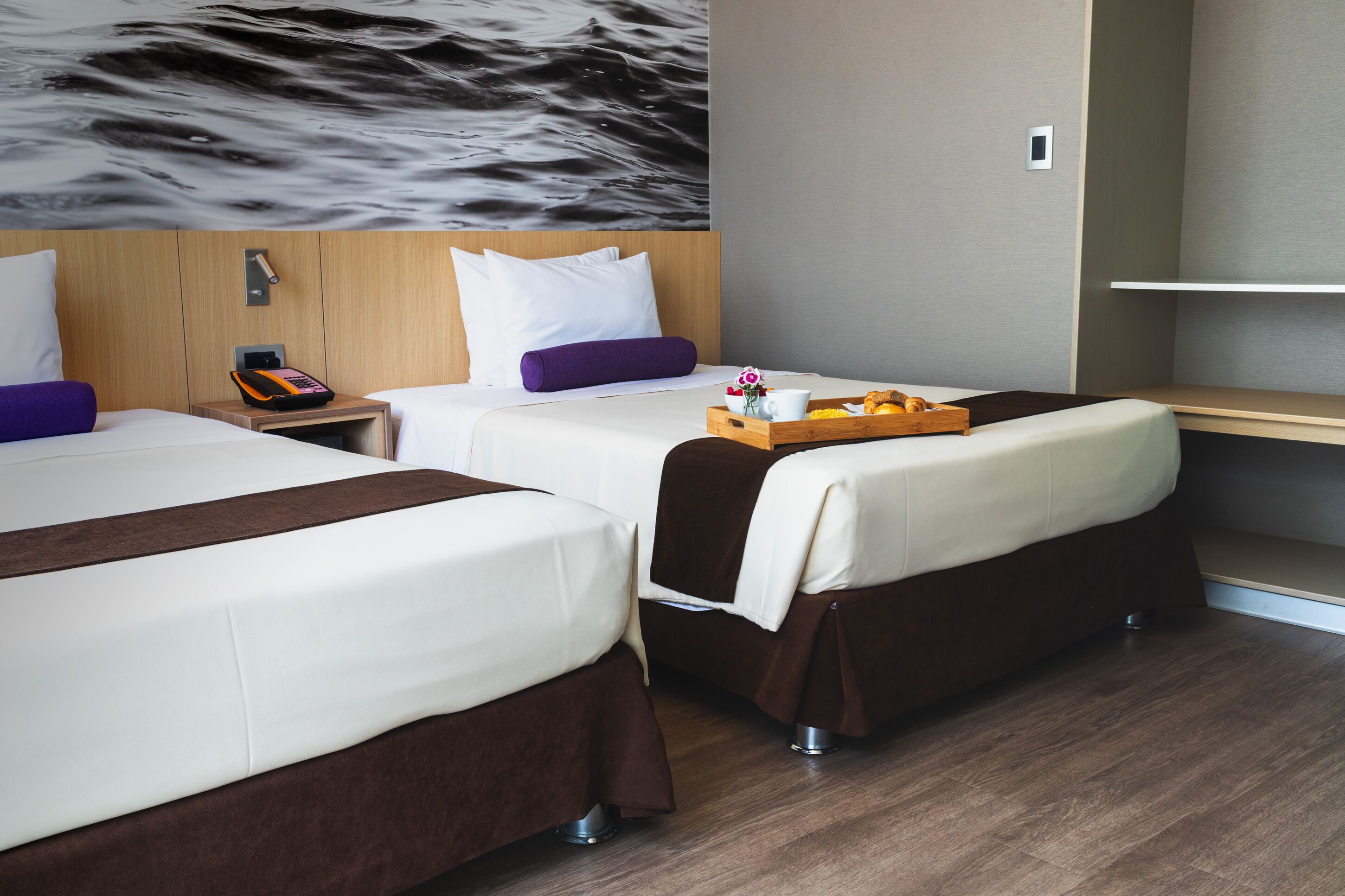 Ein Hotelzimmer mit zwei Betten, einem hölzernen Kopfteil, einem Nachttisch mit Telefon und einer Ablage für Frühstücksgegenstände sowie einem Wandbild.
