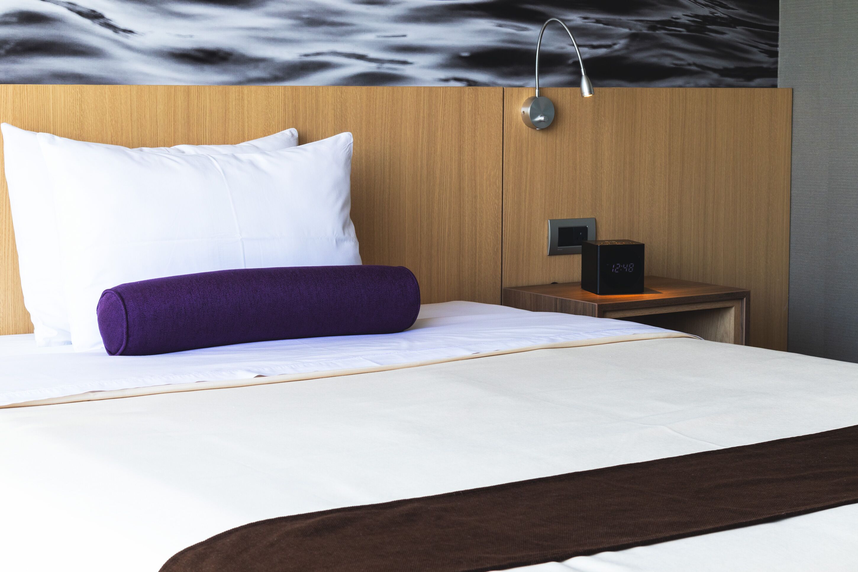 A neatly made bed with white and purple bedding, a wooden headboard, a nightstand with a clock, and a wall-mounted lamp.