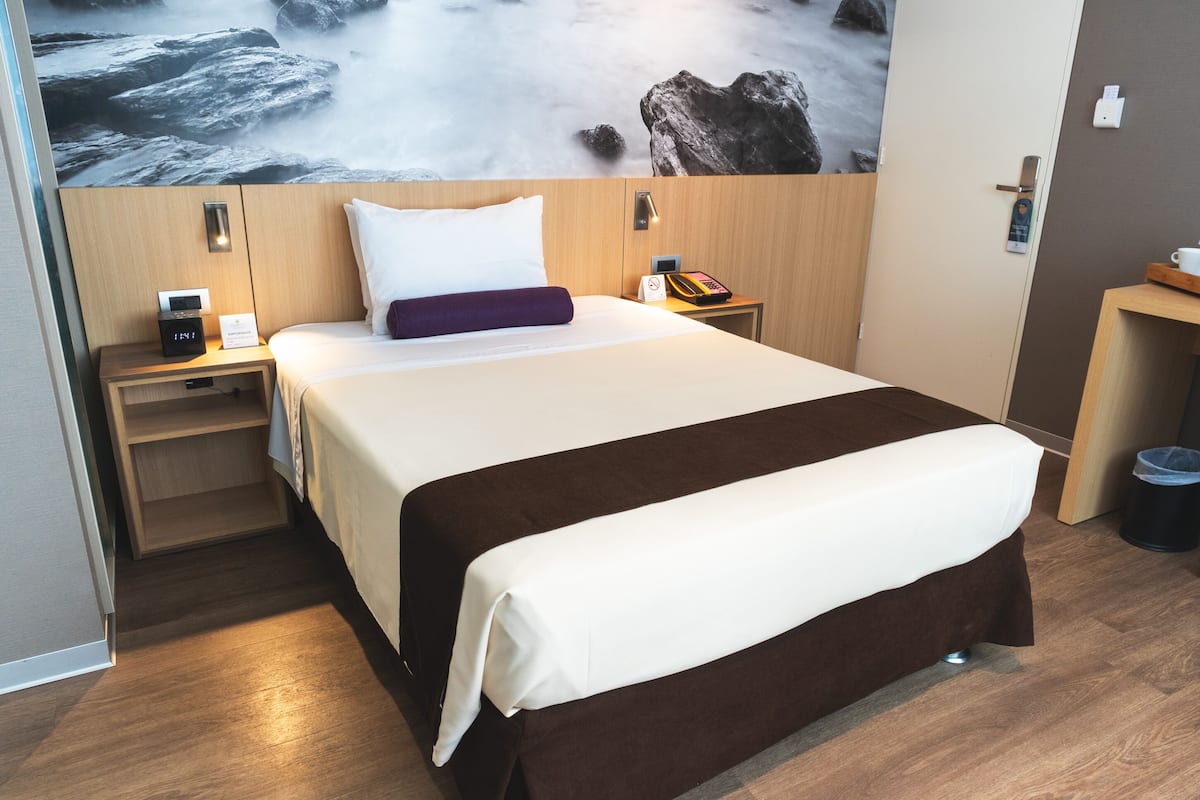 Une chambre d’hôtel avec un grand lit, une tête de lit en bois, une table de chevet avec un téléphone et une lampe, et un mur orné d’une fresque représentant un paysage.