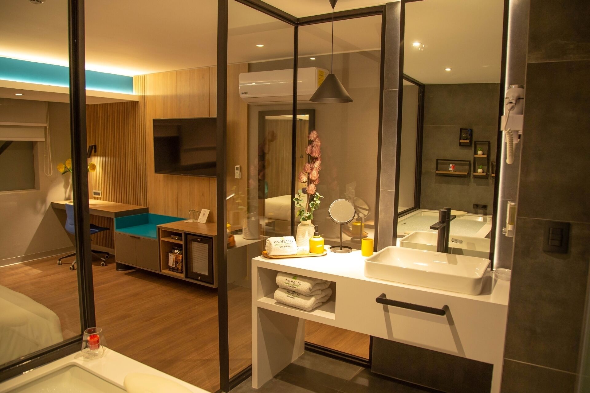 Una habitación de hotel moderna con un espejo grande, una tocador con lavabo, un televisor y un escritorio de madera con una silla.