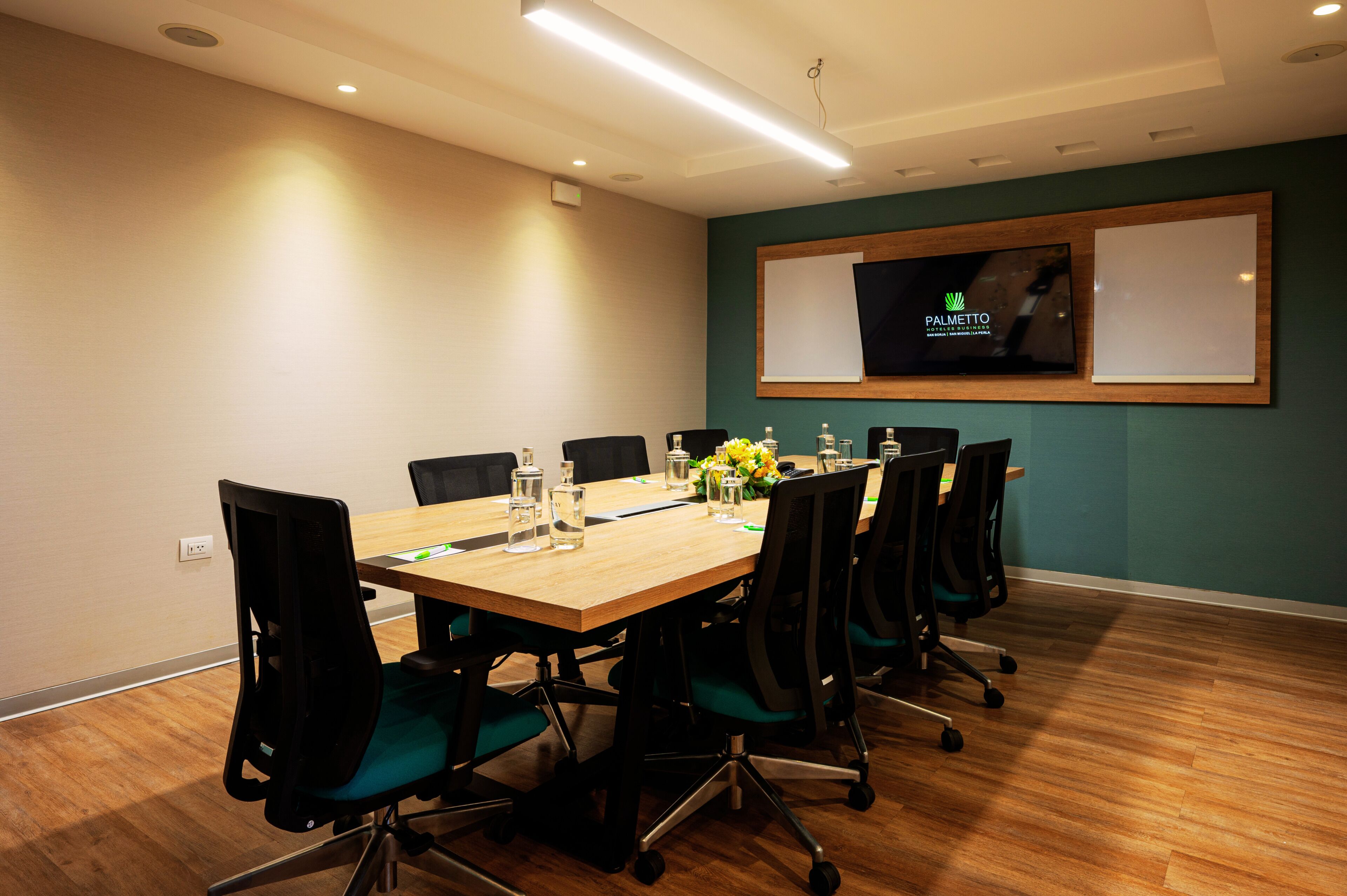 A conference room with a wooden table, black chairs, a TV screen, and a floral arrangement.