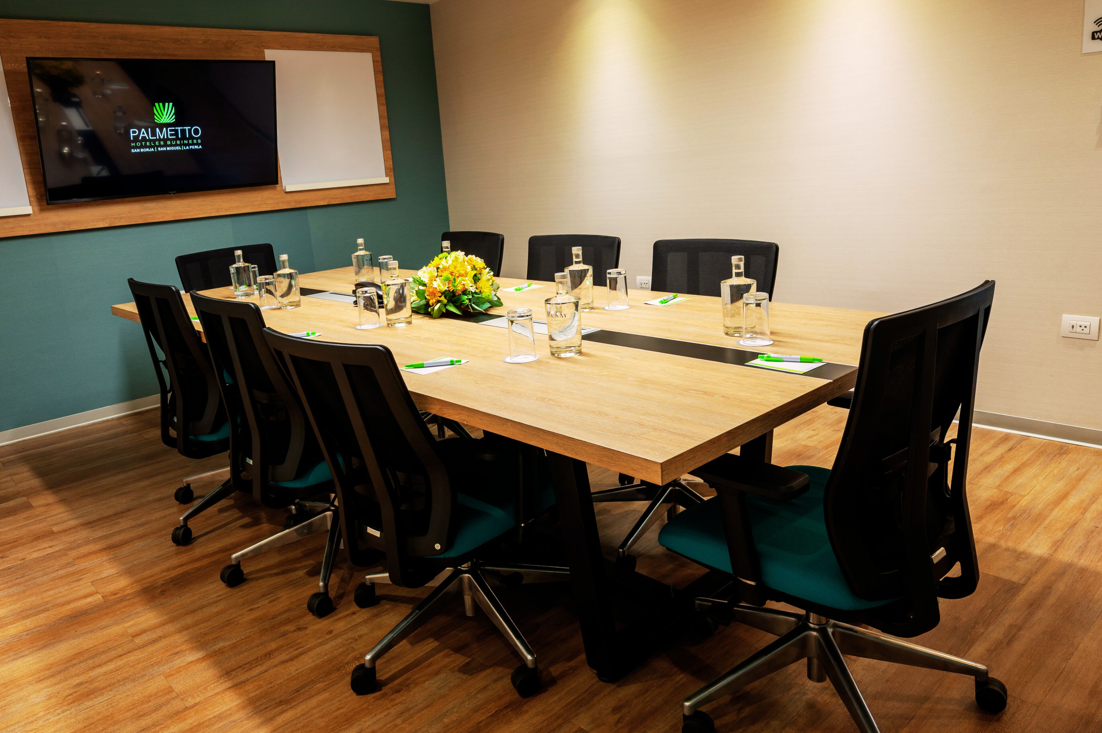 A conference room with a wooden table, black chairs, water bottles, and a floral centerpiece.