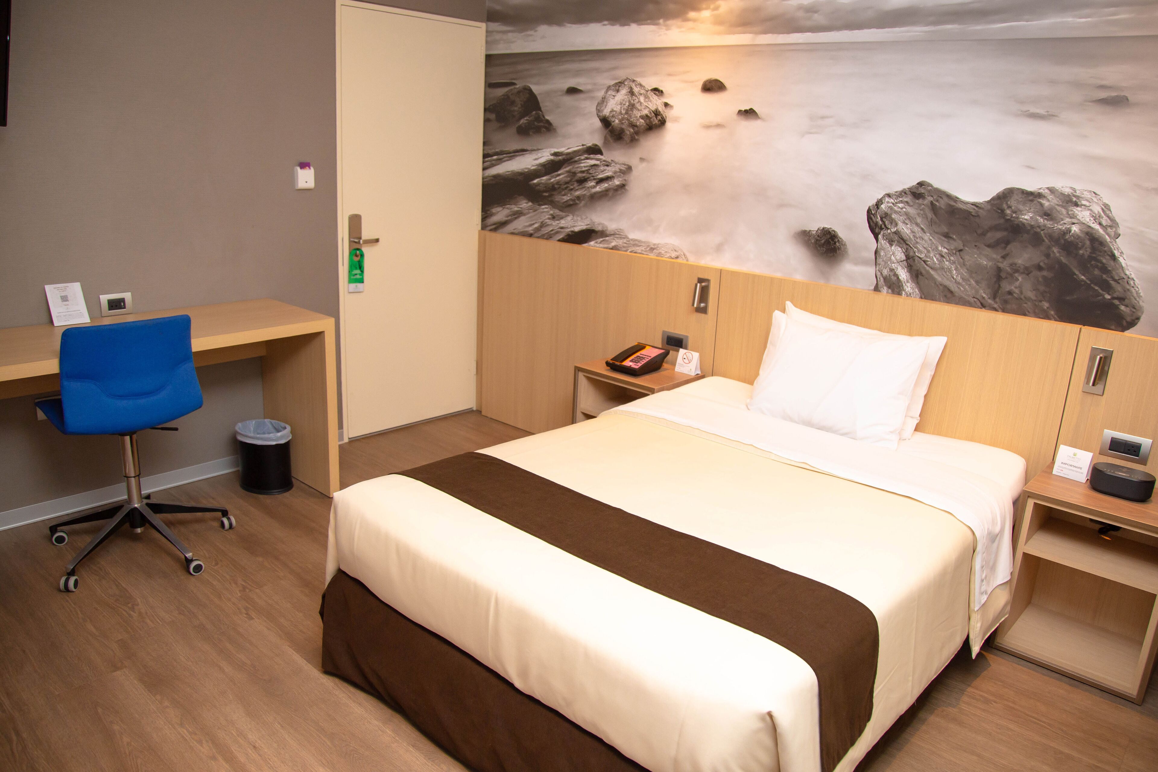 Camera d'albergo con un letto, una scrivania e una sedia. Al muro, un quadro raffigura una spiaggia.