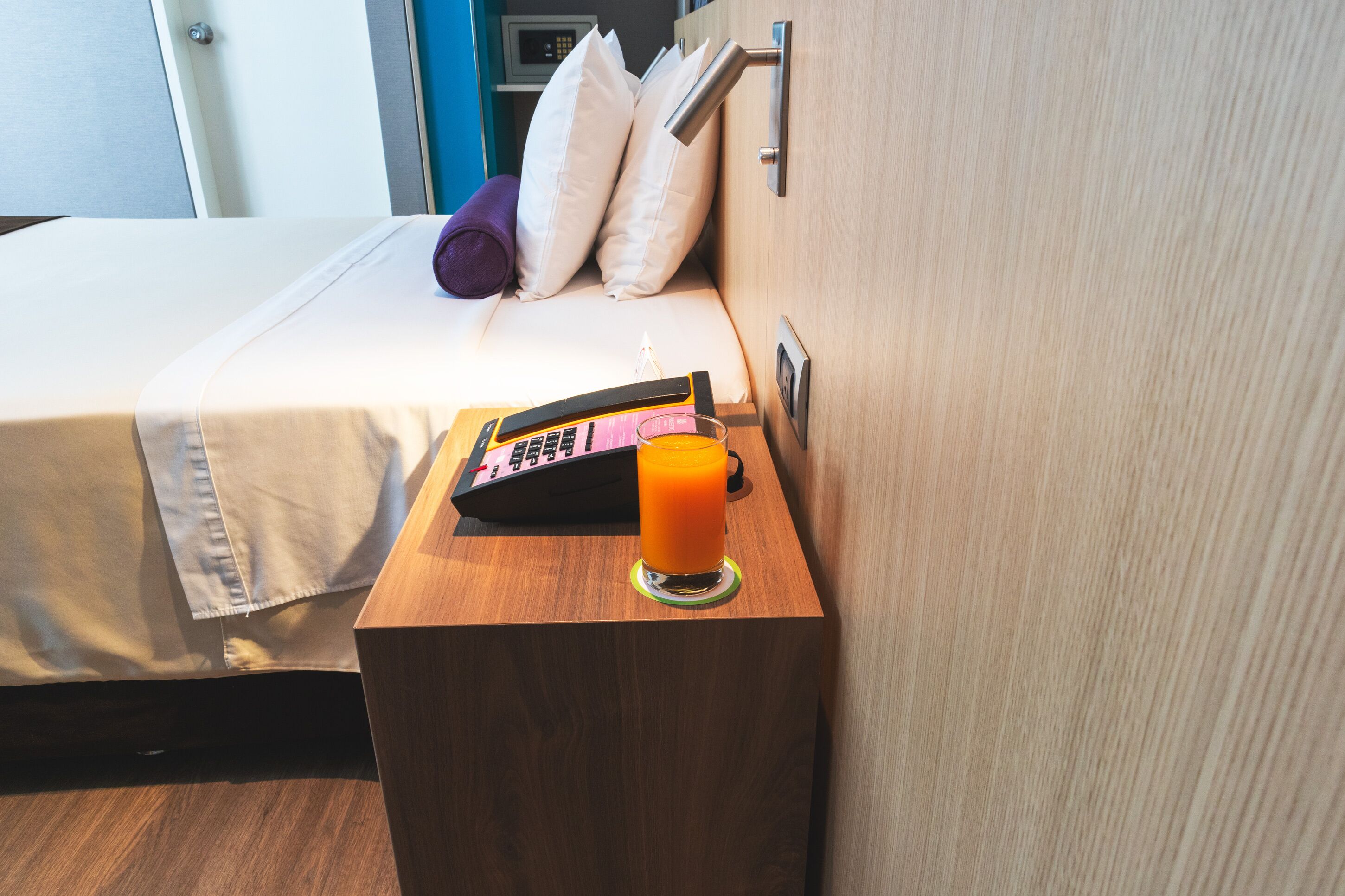 Une chambre d’hôtel avec un lit, une table de chevet sur laquelle se trouve une télécommande et un verre de jus d’orange, et une porte en bois.