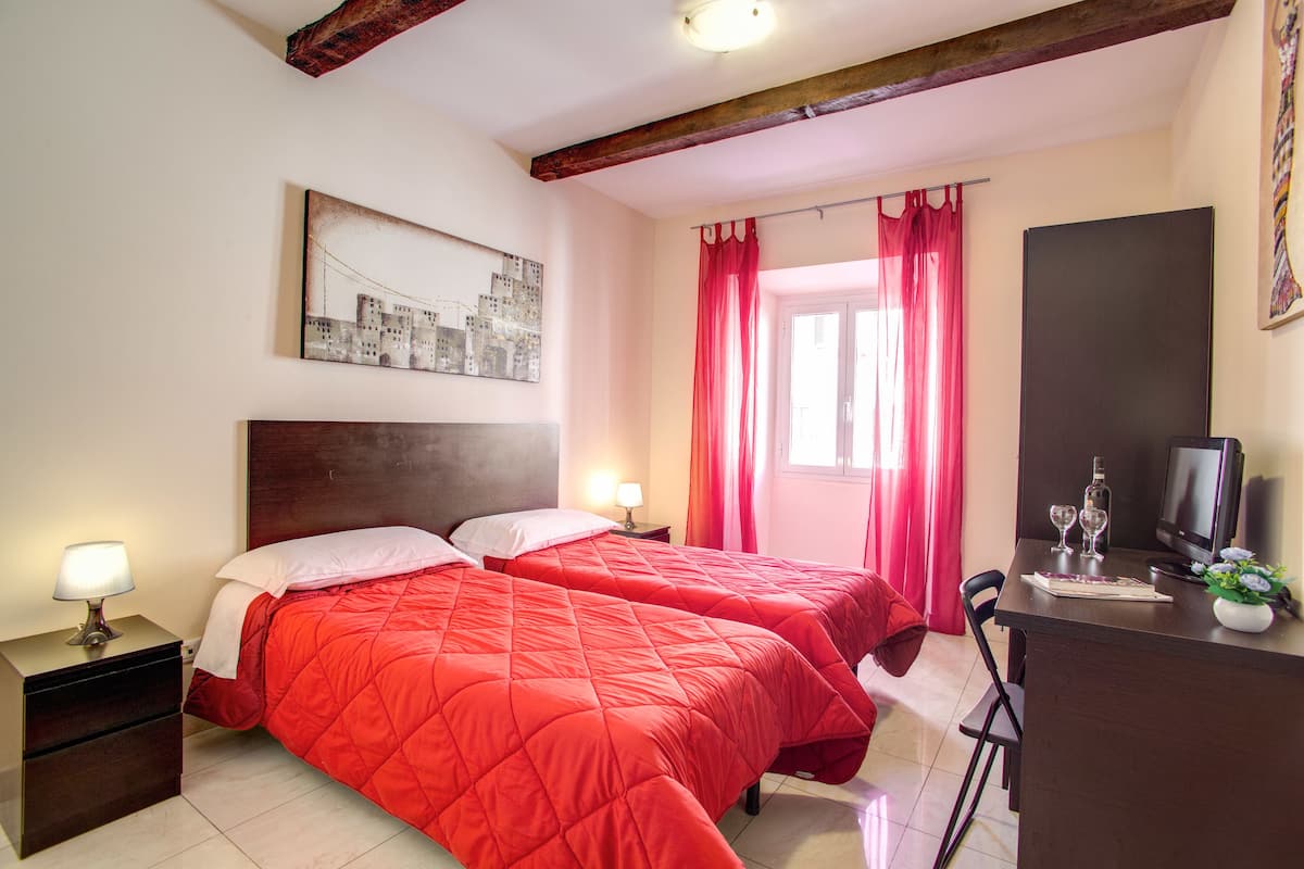 A hotel room with two beds, a desk, a TV, and a window with red curtains.