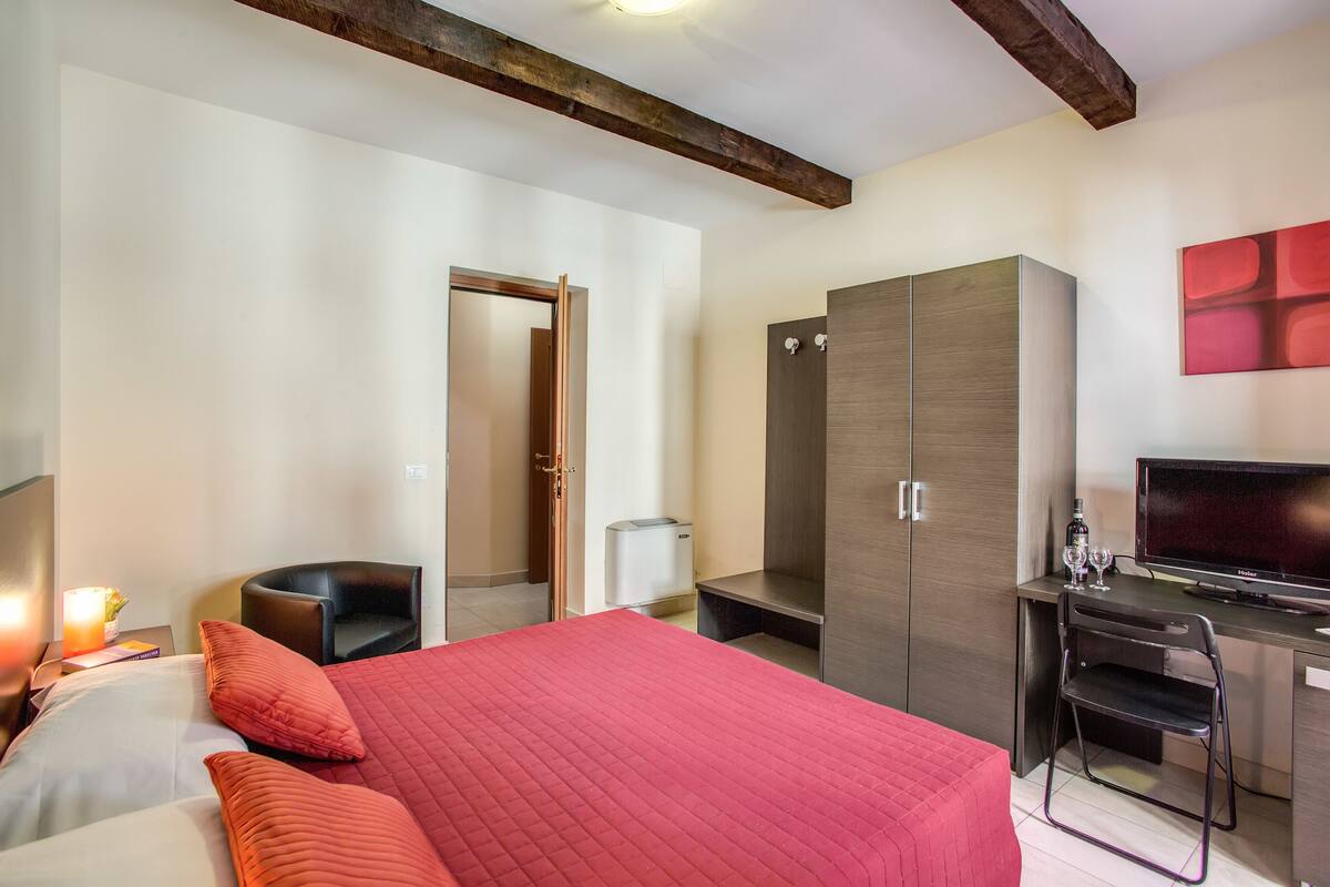 A hotel room with a large bed, a desk with a chair, a wardrobe, a television, and a small table with a bottle and glasses.