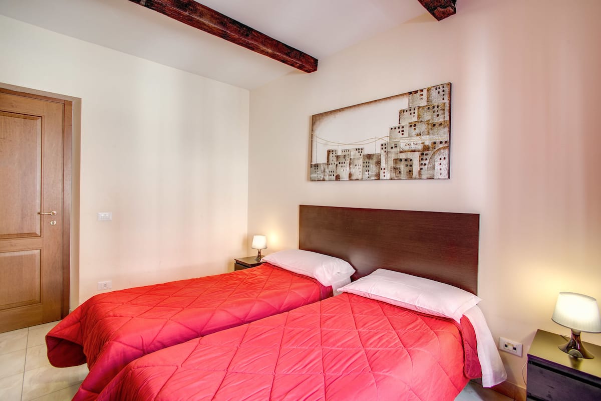 A hotel room with two beds, wooden headboards, a wall-mounted artwork, bedside lamps, and a wooden door.
