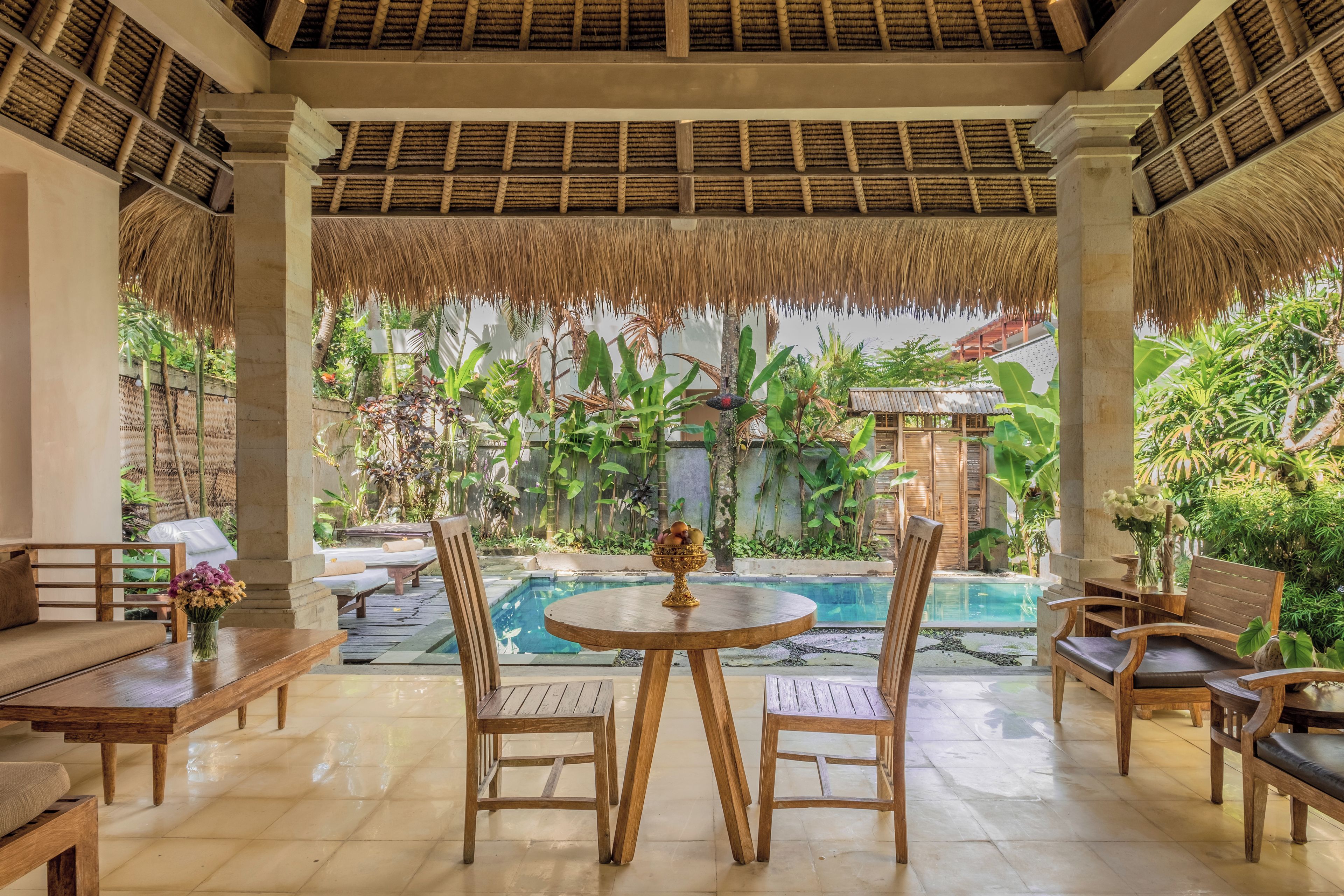 A poolside area with wooden furniture, a round table with a fruit bowl, and a view of a swimming pool and lush greenery.