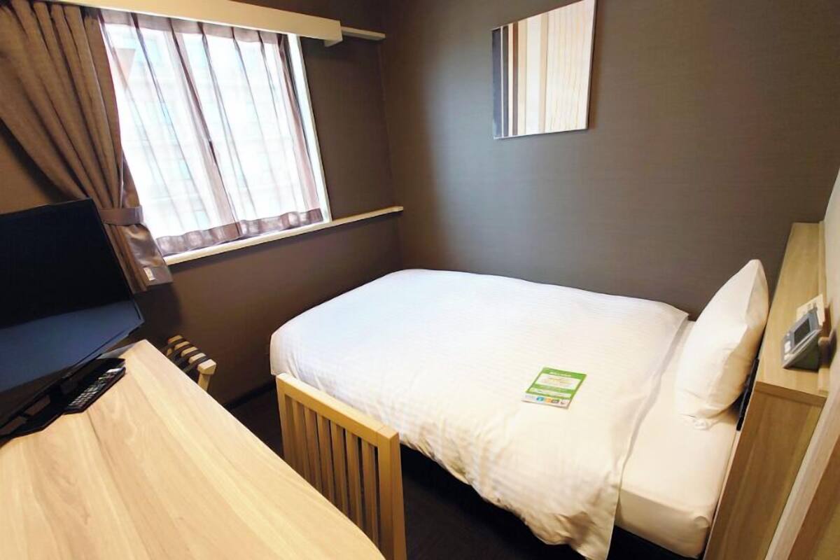 A hotel room with a single bed, a desk with a computer, a window with curtains, and a small bedside table with a lamp.