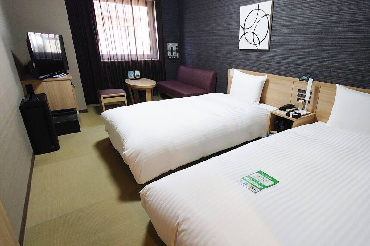 A hotel room with two beds, a desk with a computer, a chair, a TV, and a window with curtains.