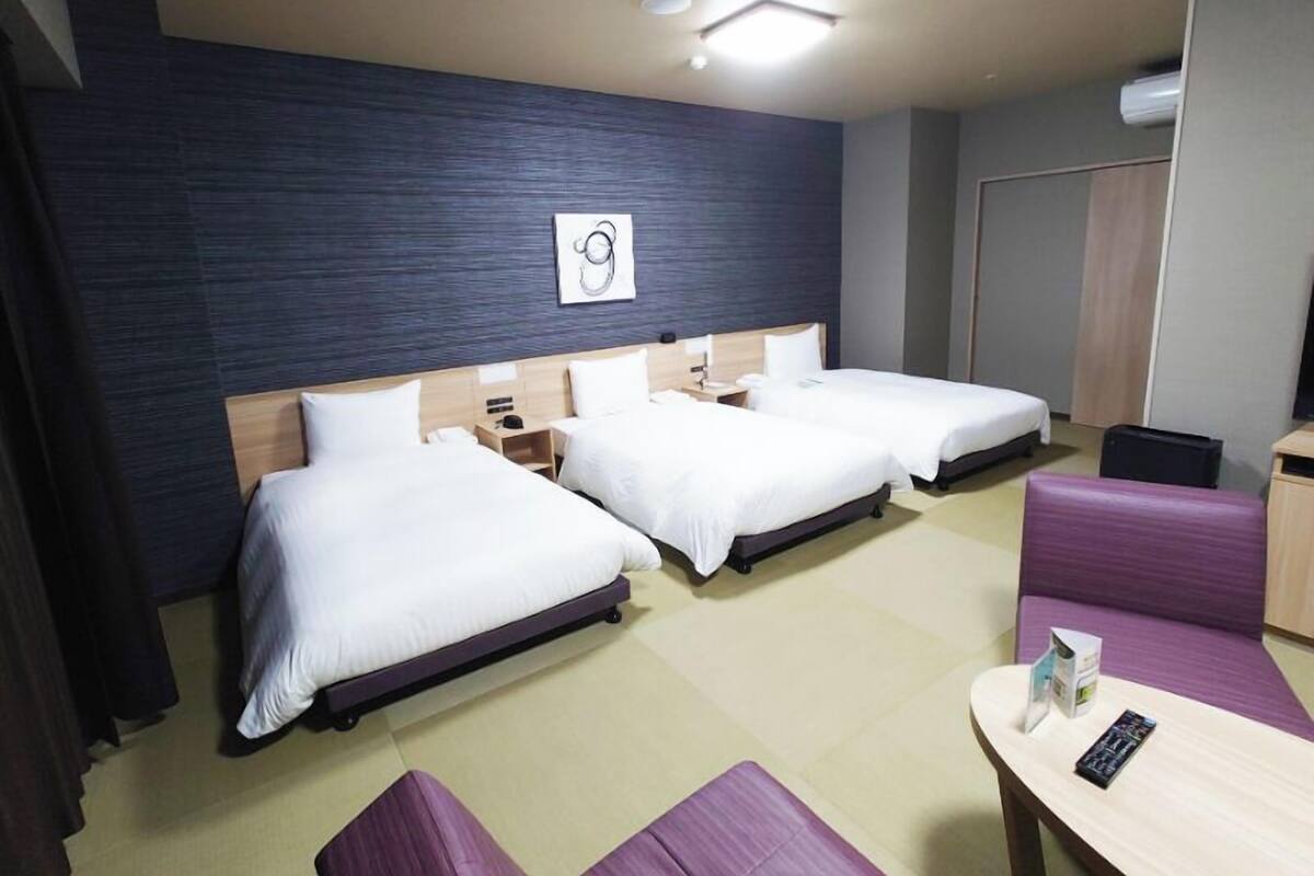 A hotel room with three beds, a small table, and a purple sofa.