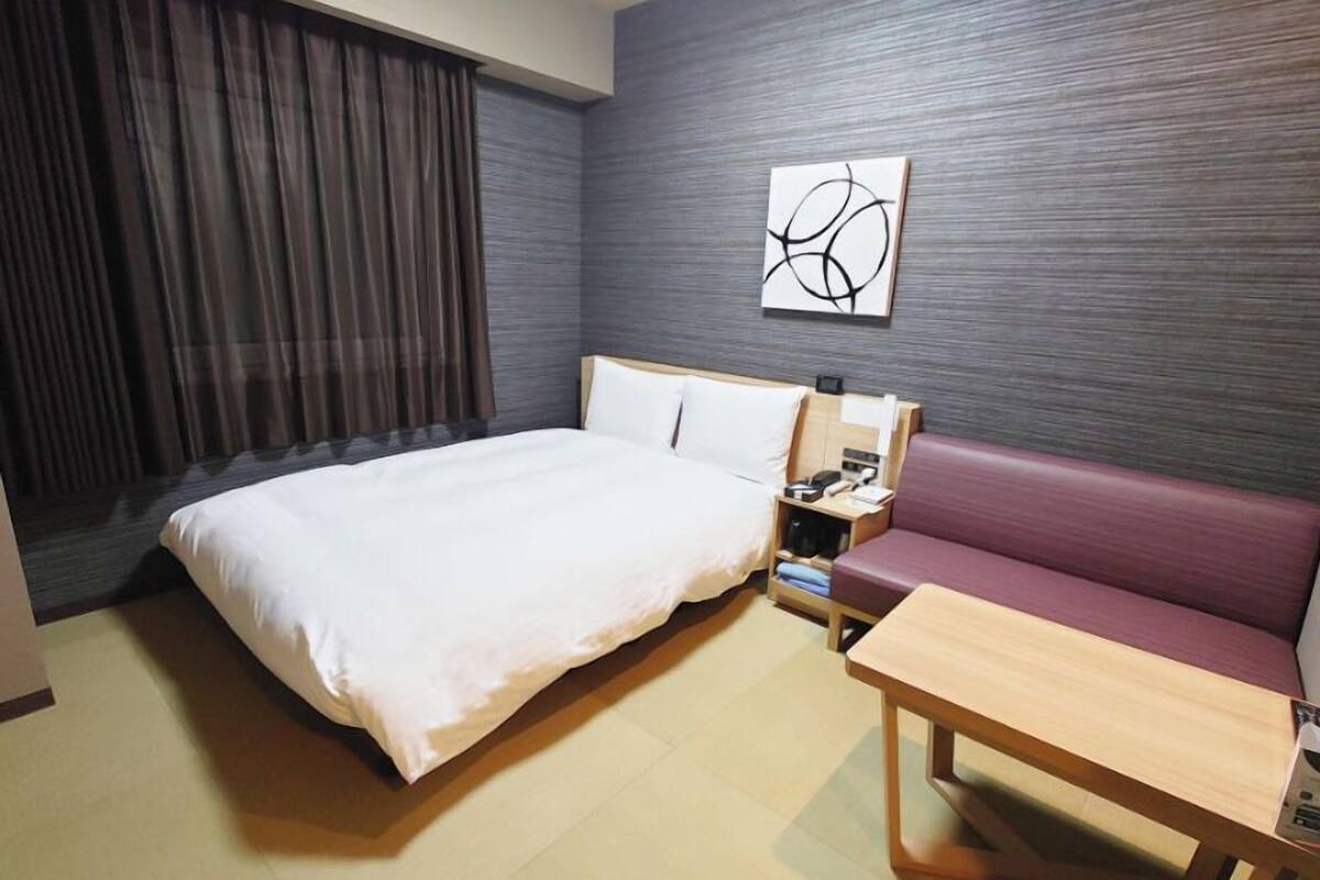 A hotel room with a bed, a wooden nightstand, a table, a sofa, and a wall art piece.