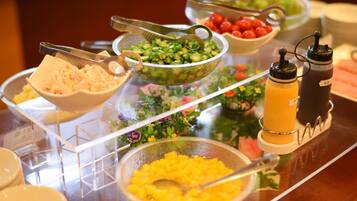 A buffet with various food items including shredded cheese, green vegetables, tomatoes, and a yellow drink.