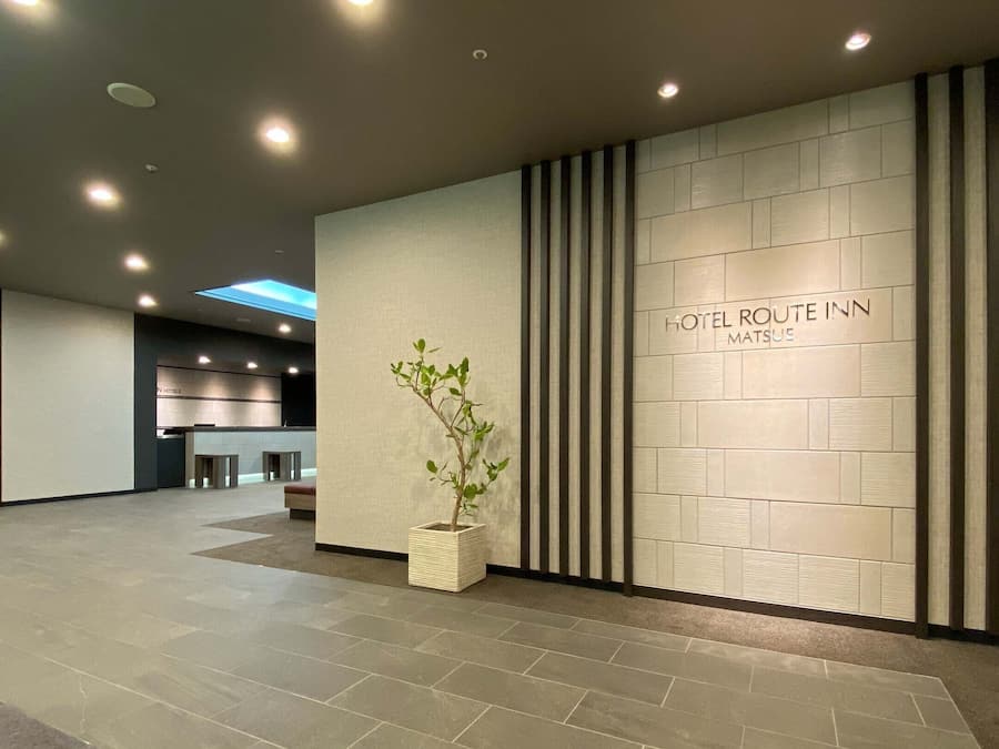 A modern hotel reception area with a reception desk, a potted plant, and a wall with the hotel name.