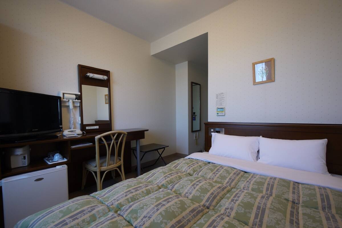 A hotel room with a bed, a desk with a television, a chair, a small refrigerator, and a mirror.