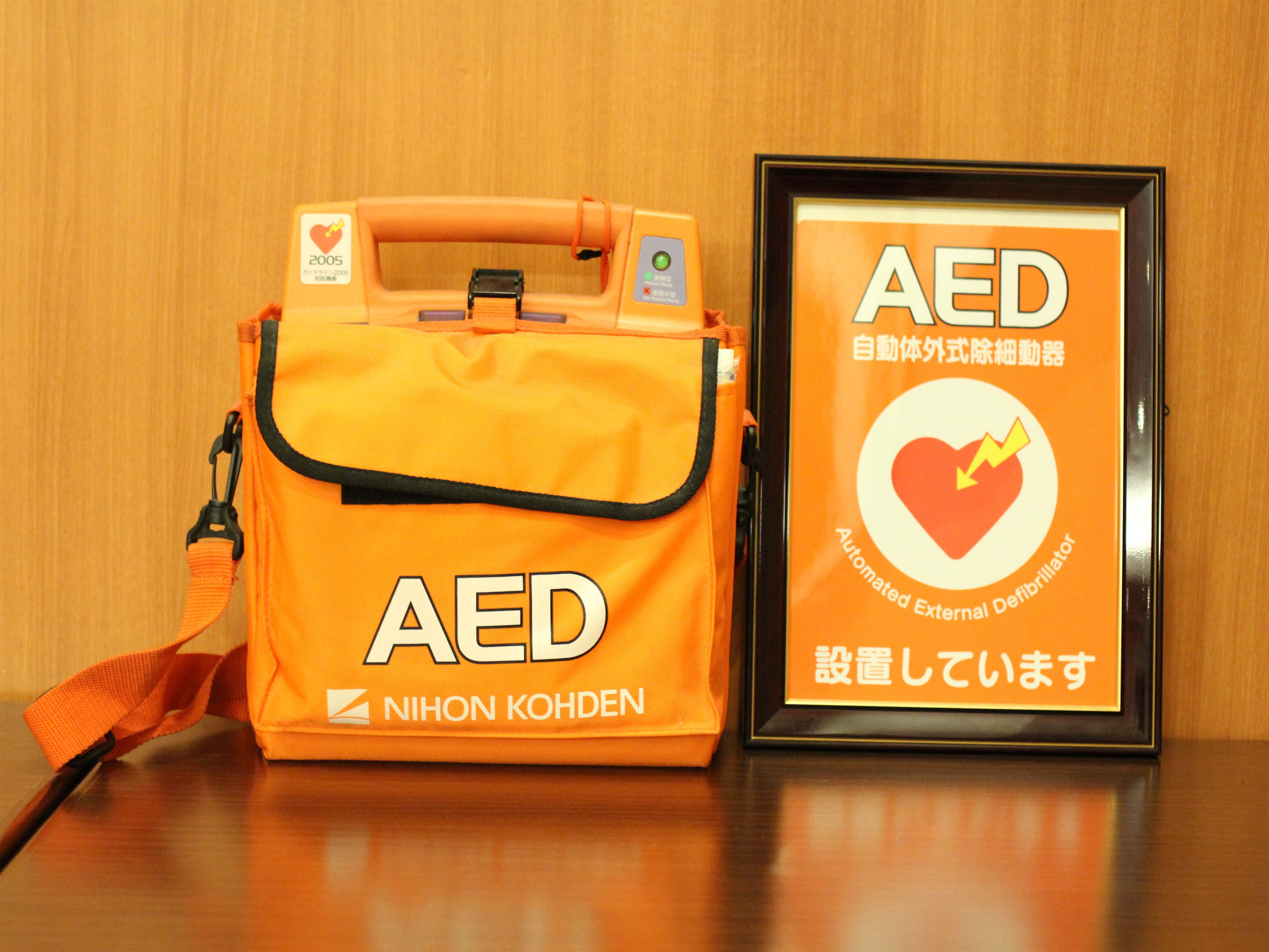Eine AED-Tasche und ein gerahmtes Schild mit AED-Hinweis auf einer Holzoberfläche.
