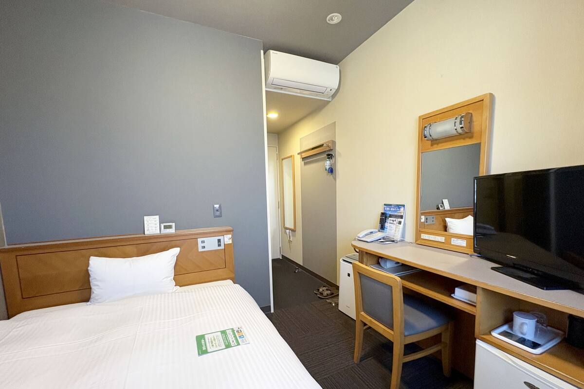 A hotel room with a bed, a desk with a television, a mirror, and a small refrigerator.