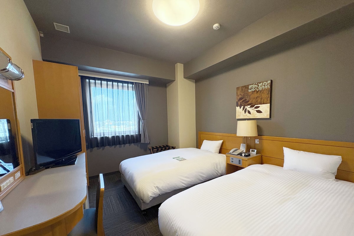 A hotel room with two beds, a desk, a television, a window with curtains, a lamp, and a wall-mounted artwork.