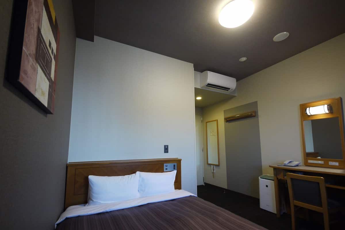 A hotel room with a bed, a desk, a chair, a mirror, an air conditioning unit, and a door leading to another room.