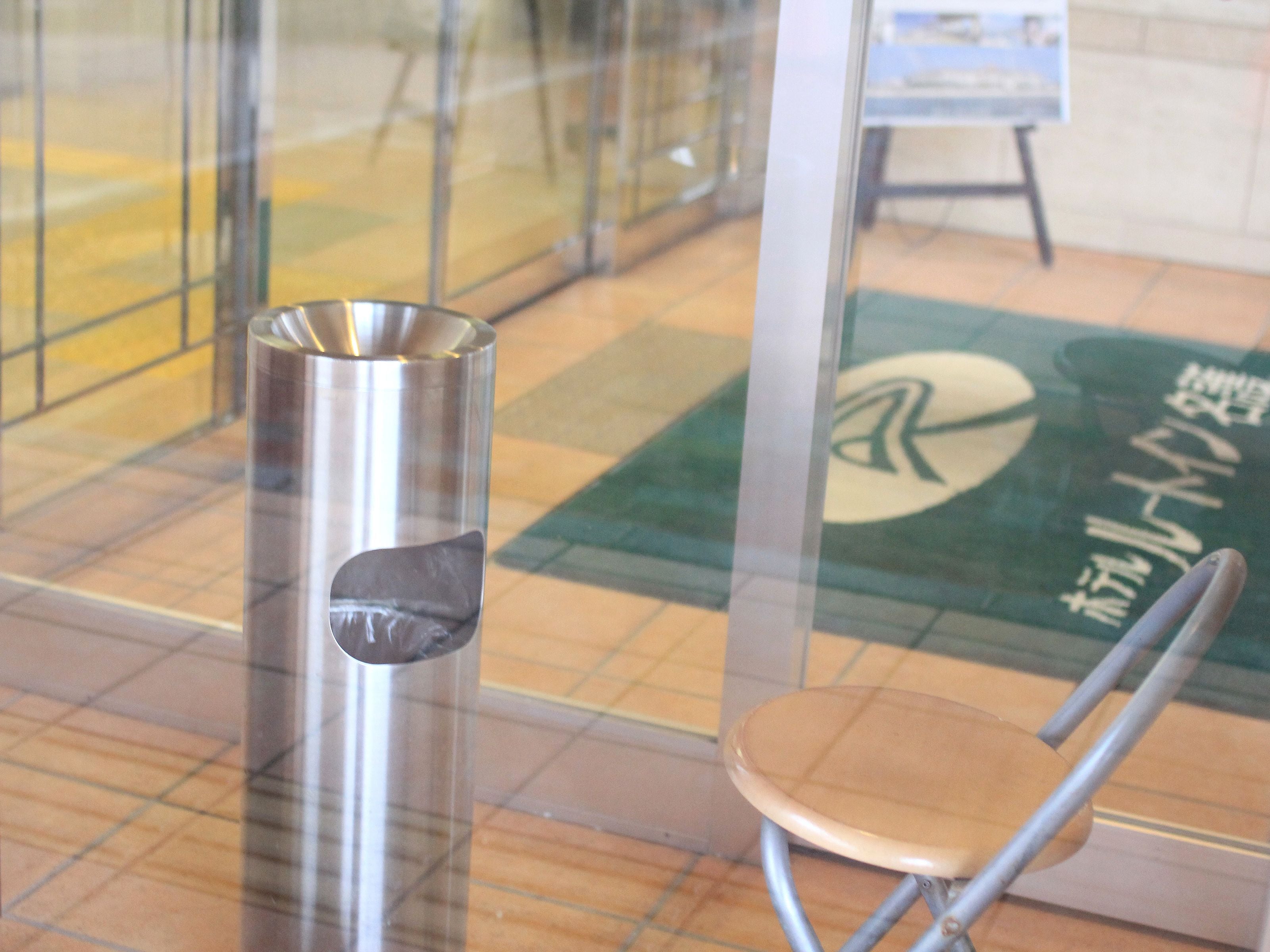 A metallic trash can and a chair in a public indoor space with tiled flooring and a glass partition.