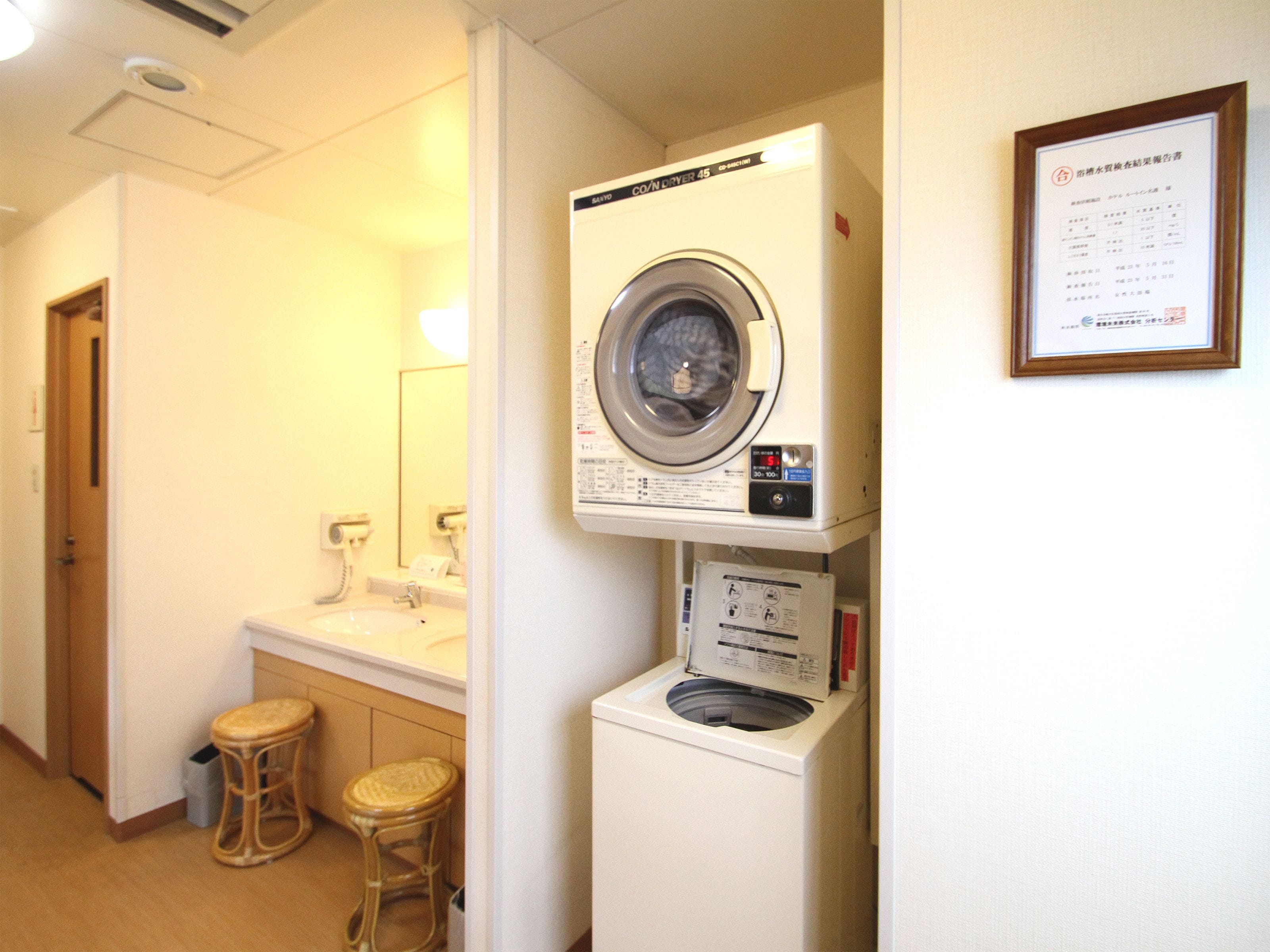 A laundry room with a front-loading washing machine, a dryer, and a sink. There are two stools and a trash can.