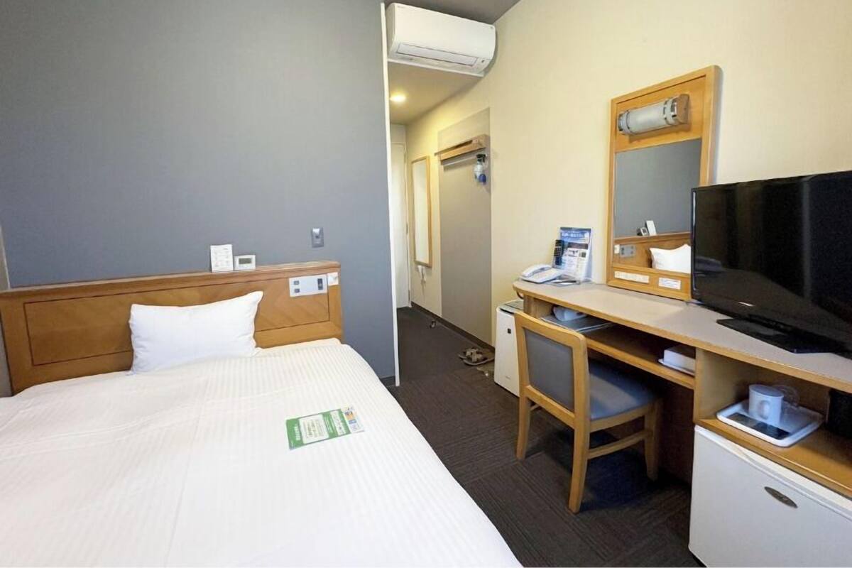 A hotel room with a bed, a desk with a chair, a television, and an air conditioning unit.
