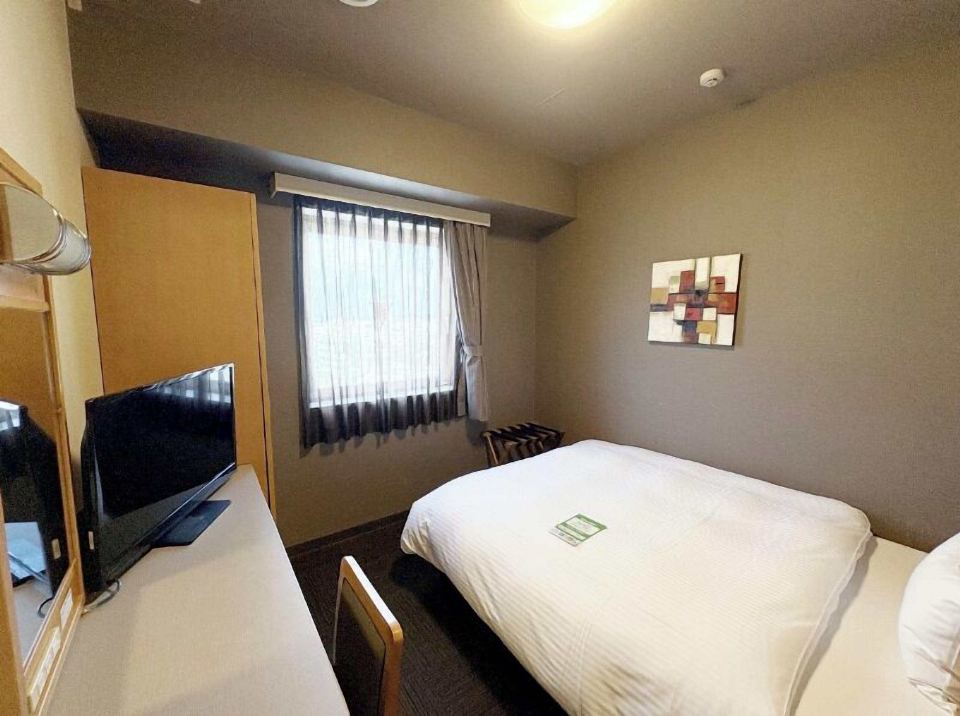 Ein Hotelzimmer mit zwei Betten, einem Schreibtisch, einem Flachbildfernseher, einem Fenster mit Vorhängen und einem an der Wand befestigten abstrakten Kunstwerk.