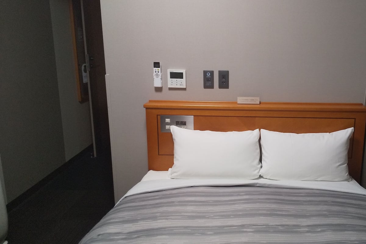 A hotel room with a wooden headboard, a bed with white and gray striped bedding, and a control panel with a thermostat and a lock on the wall.