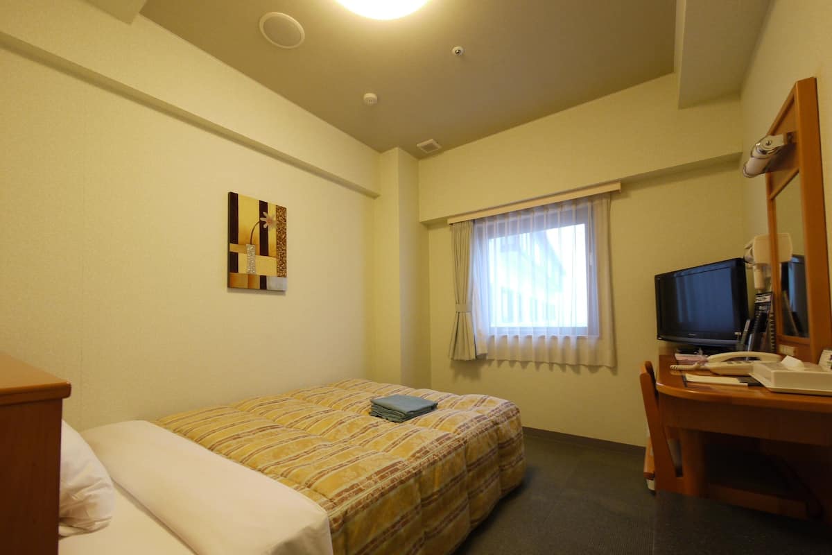 A hotel room with a bed, a desk with a computer, a chair, a window with curtains, and a small painting on the wall.
