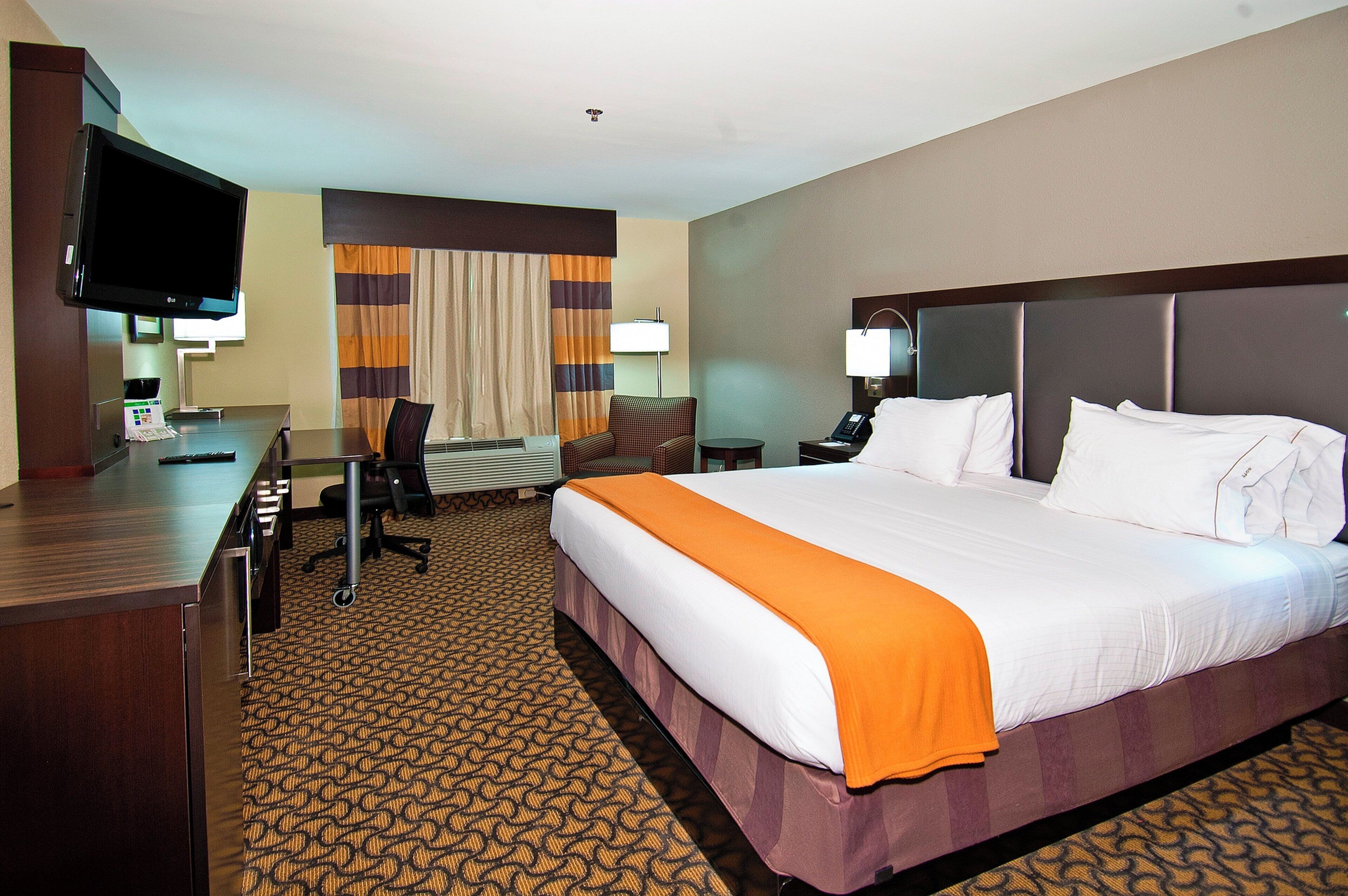 Quarto de hotel com uma cama grande, escrivaninha, cadeira, televisão e janela com cortinas.