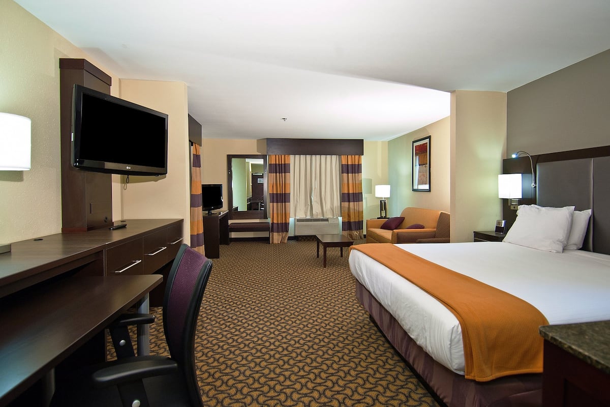 A hotel room with a bed, desk, chair, television, and a view of the hallway.